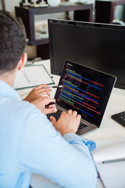 A person wearing a light blue shirt is seated at a desk with their back to the camera, working on a laptop computer displaying lines of colorful code or script on the screen. The workspace includes a 