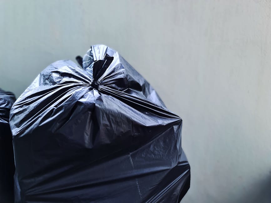 A large collection of mixed household waste and recyclables, including cardboard boxes, plastic bags, and paper waste, is piled around a communal rubbish bin situated on a paved parking area in an urb