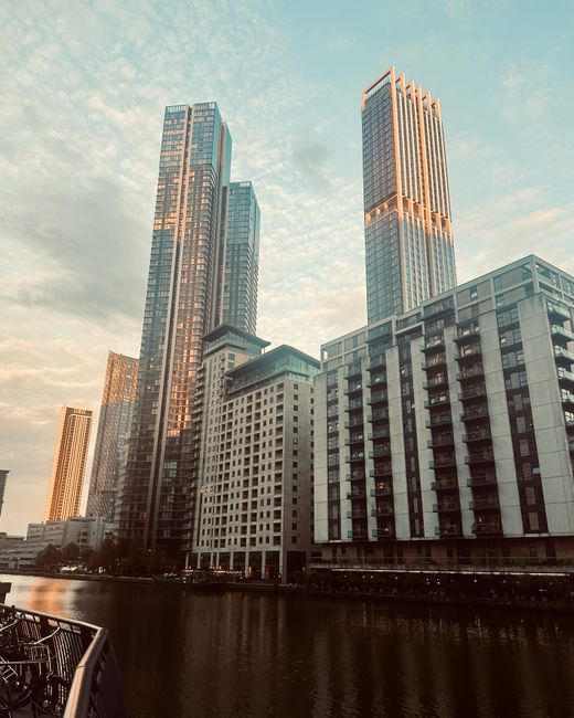 A nighttime cityscape featuring a cluster of illuminated high-rise office buildings along a river, with reflective water in the foreground. The skyscrapers have glass facades with bright lights in var
